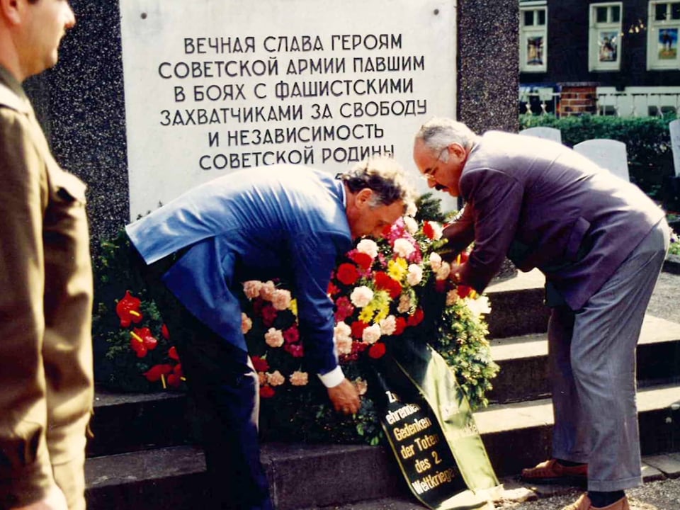 Zwei Männer legen eine Blumenkranz nieder auf einem Grab. Grabstein mit kyrillischer Schrift.