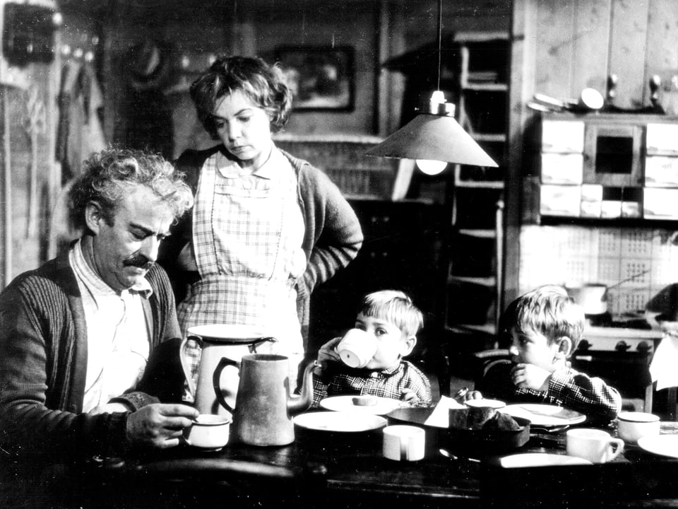 Eine Familie am Esstisch. Die Mutter mit Schürze hat die Hände in die Hüften gestemmt und steht neben ihrem Mann. Zwei kleine Jungen sitzen neben ihm am Tisch.