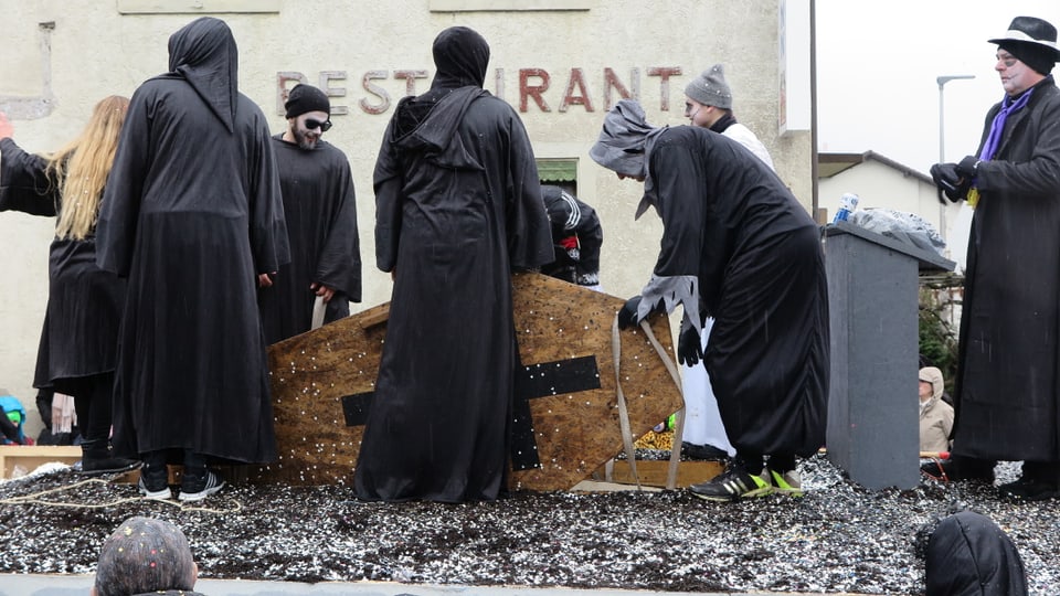 Schwarz gekleidete Figuren versenken einen Sarg in einem Boden aus Konfetti