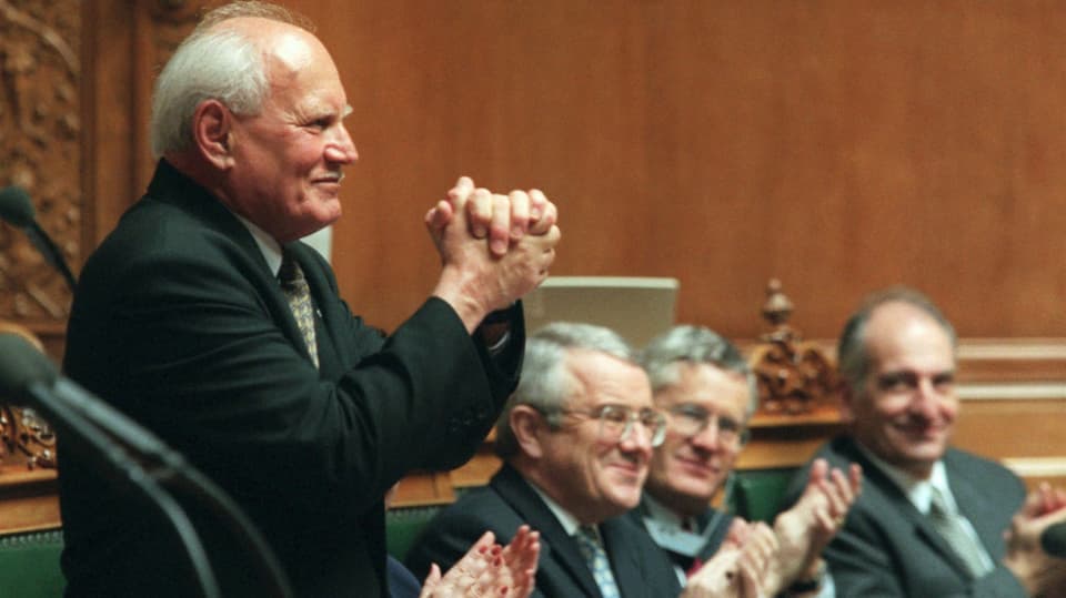 The man raises his hands in thanks, applauding members of parliament in the background.