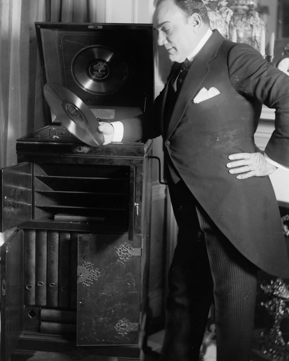 Ein Mann im Anzug steht neben einem alten Schaltplattenspieler und hält eine Platte in der Hand.