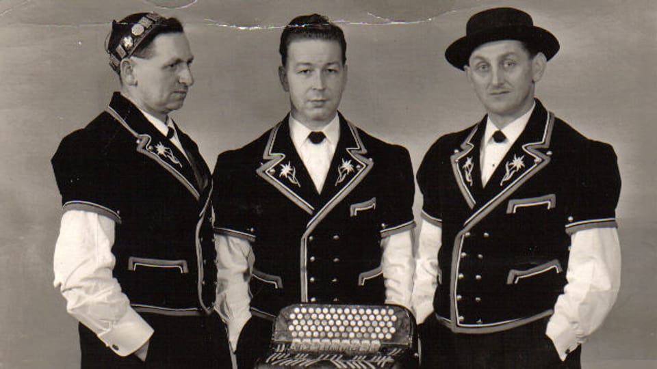 Schwarz-weiss Fotografie mit drei Jodlern in Sennentracht vor einem Akkordeon.