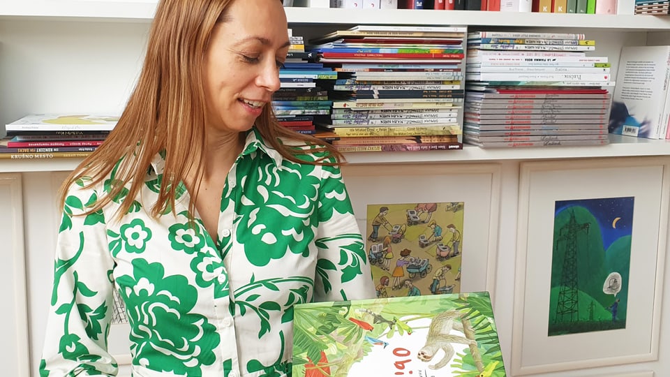 Woman with long light brown hair and a blouse with a green floral pattern holds a book in her hand
