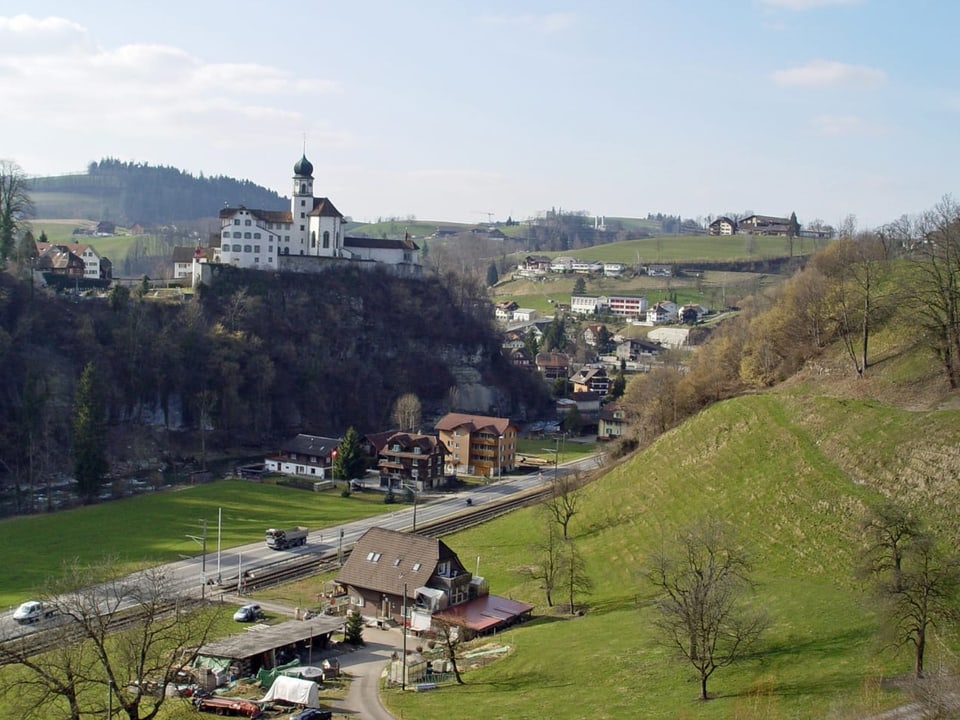 Strassendorf inmitten grüner Hügel, darüber throhnt ein Barockkloster