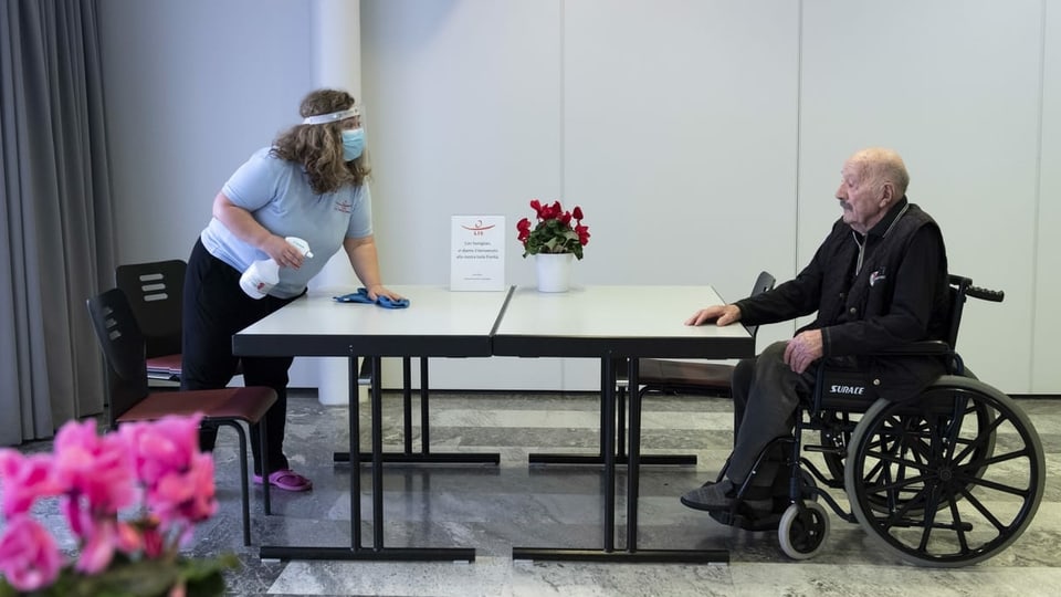 Eine Frau reinigt einen Tisch, ein älterer Mann sitzt im Rollstuhl.