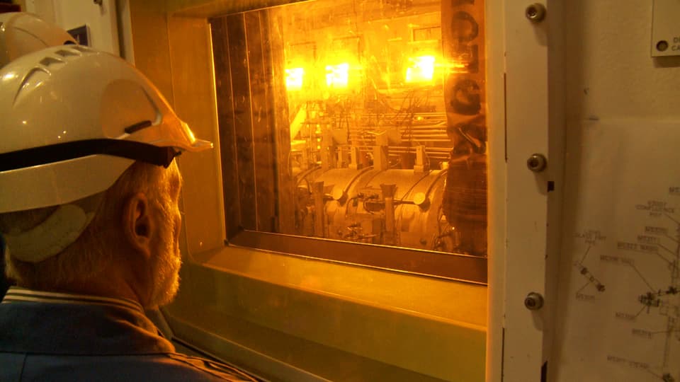 Ein Mann mit Schutzhelm blickt durch ein Fenster auf ein hell beleuchtetes Gerät