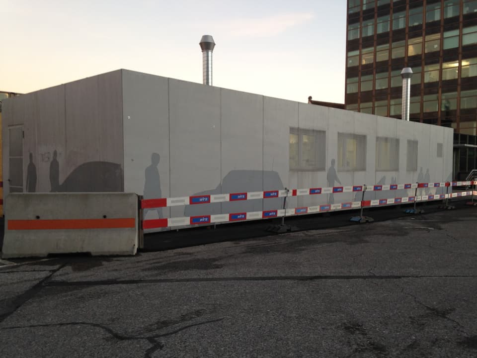 Grauer Container mit Bauabschrankung, auf dem Container sind Silhoutten von Menschen und Autos aufgezeichnet.