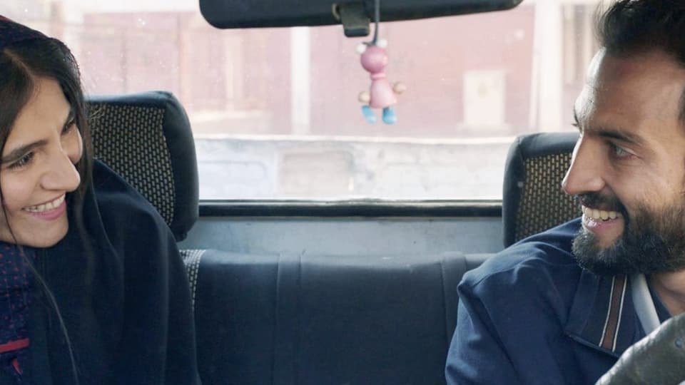 Filmszene: Eine Frau und ein Mann sitzen auf den Vordersitzen eines Autos und lächeln sich an.