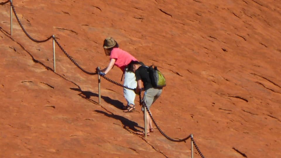 Eine Frau in pinkigem Shirt und ein Mann mit dunklem Shirt halten sich an einer Kette und klettern auf den Berg.