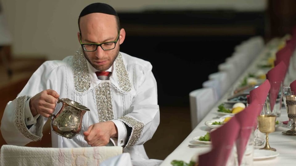 Ein jüdischer Rabbi sitzt an einer langen gedeckten Tafel und hält eine silbrige Kanne in der Hand.