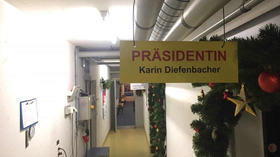 Ein langer Gang in einem Zivilschutzbunker. Im Vordergrund ein gelbes Schild mit der Aufschrift: Präsidentin. Karin Diefenbacher.