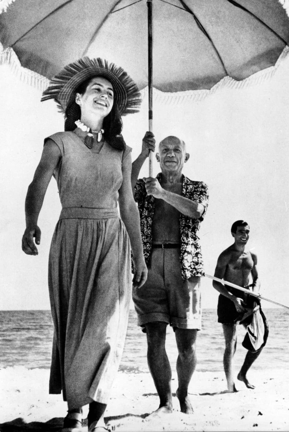 Ein stehender Mann am Strand hält einen grossen Sonnenschirm über eine junge lächelnde Frau mit Strohhut.