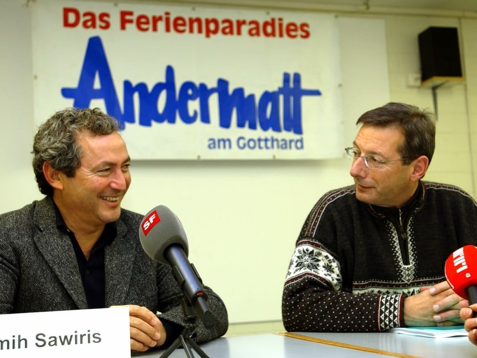 Zwei Männer lächeln sich an. Vor ihnen stehen Mikrofone, hinter ihnen ein Plakat mit der Aufschrift "Das Ferienparadies Andermatt am Gotthard".