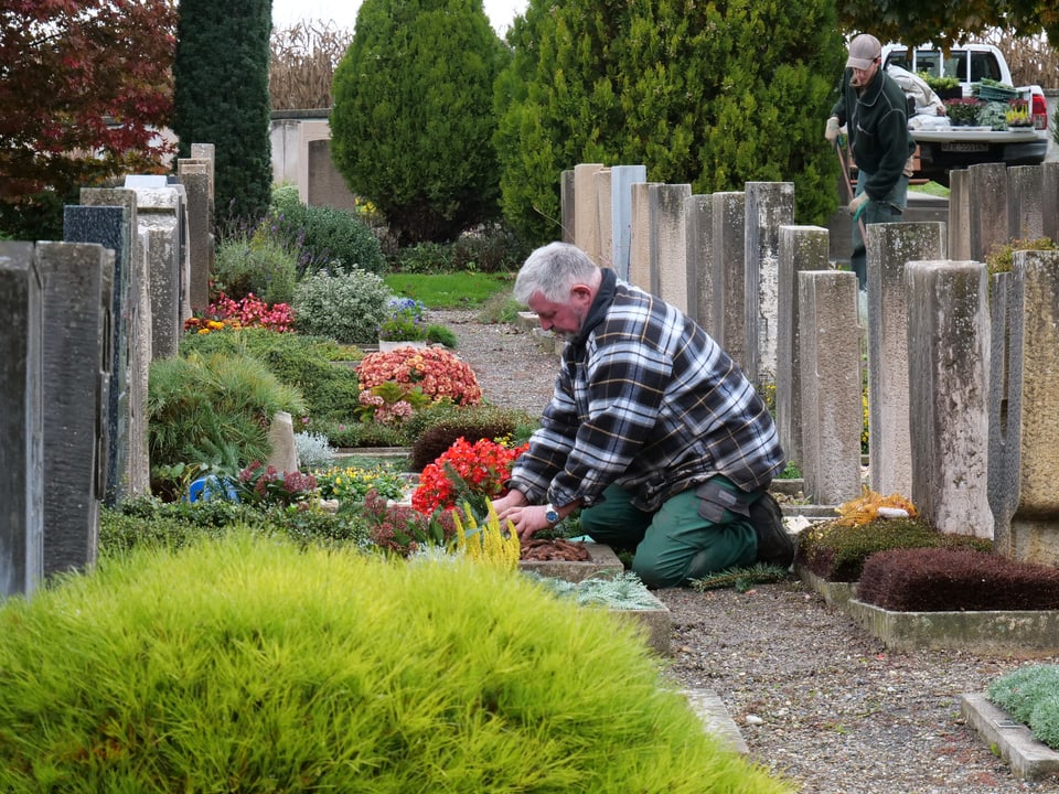 Ein Friedhof mit verschiedenen Gräbern, Pflanzen, zwei Männer am Arbeiten