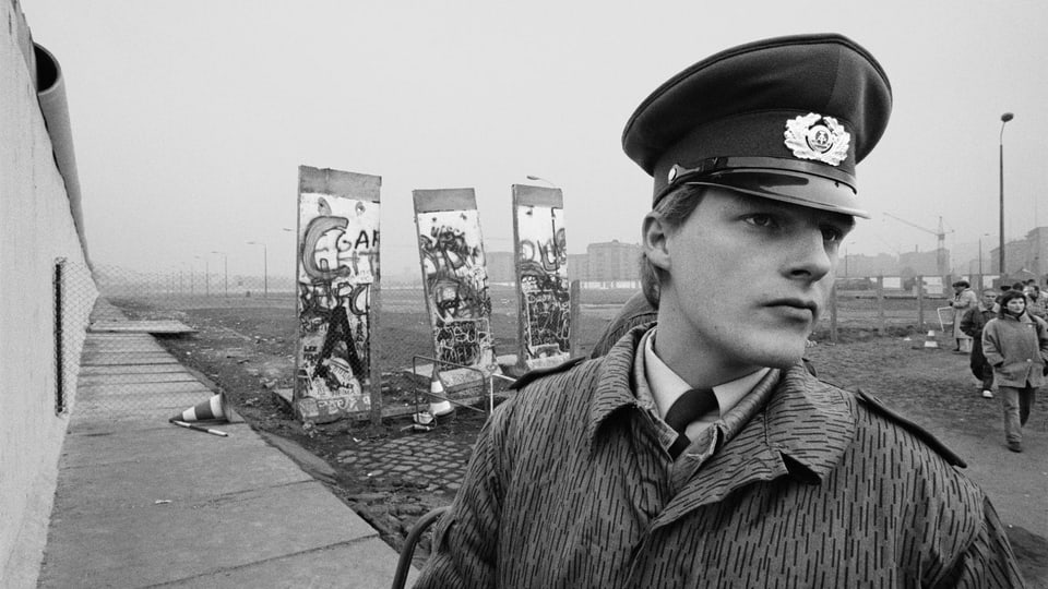 Eine Schwarz-Weiss Aufnahme eines Polizisten, mit Überresten der Berliner Mauer