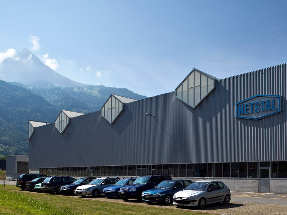 Fabrikhalle der Maschinenfabrik Netstal mit parkierten Autos davor, im Hintergrund die Glaner Berge