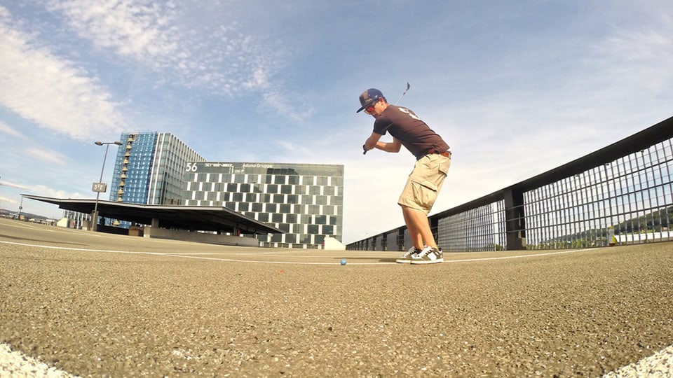 Mann spielt Golf auf einem urbanen Platz mit modernen Gebäuden im Hintergrund.