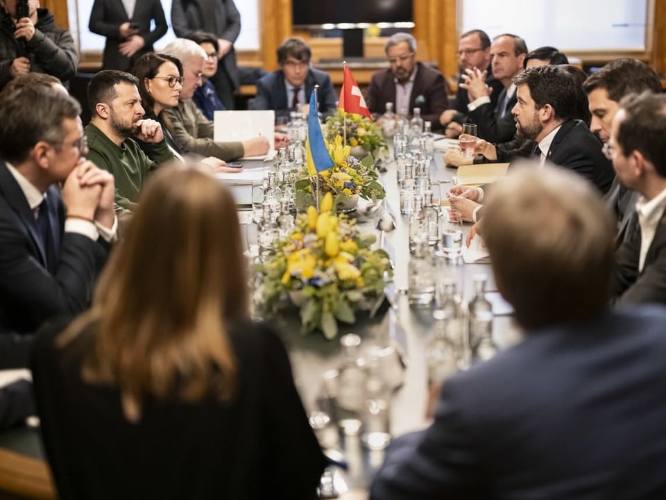 Parlamentarier sitzen um ovalen Tisch mit Schweizer- und Ukraine-Fahne.