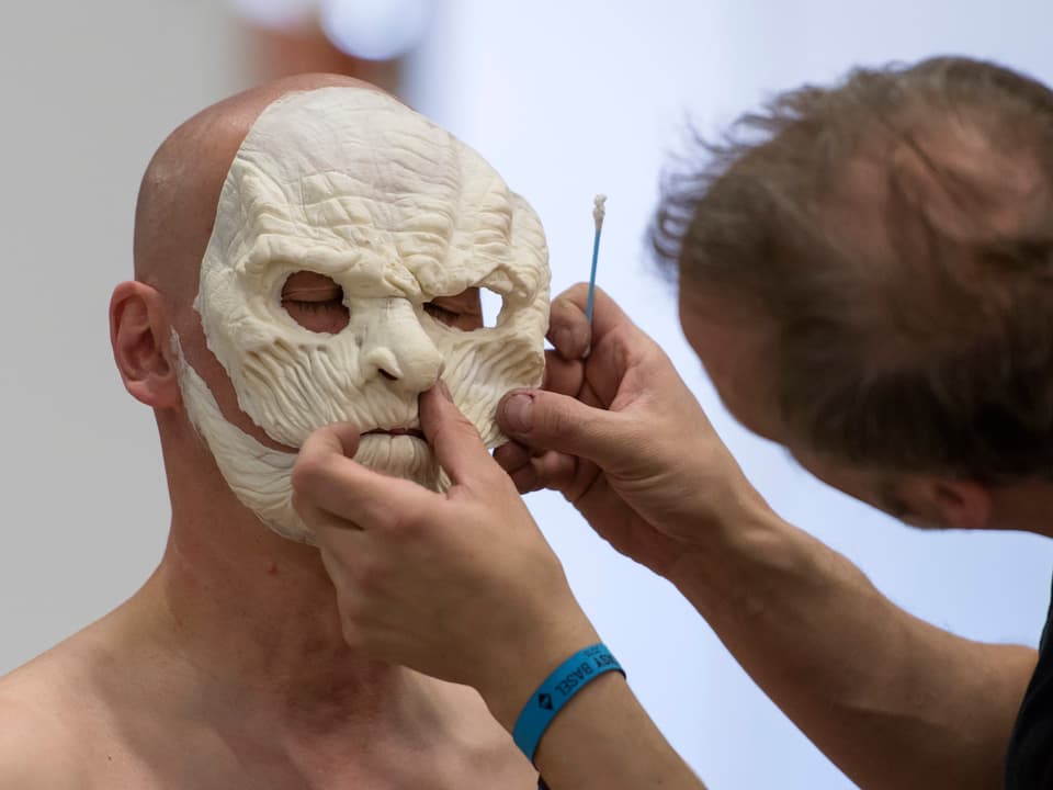 Ein Make-up-Artist bei der Arbeit. Er setzt einem Mann einen Teil einer Maske auf.