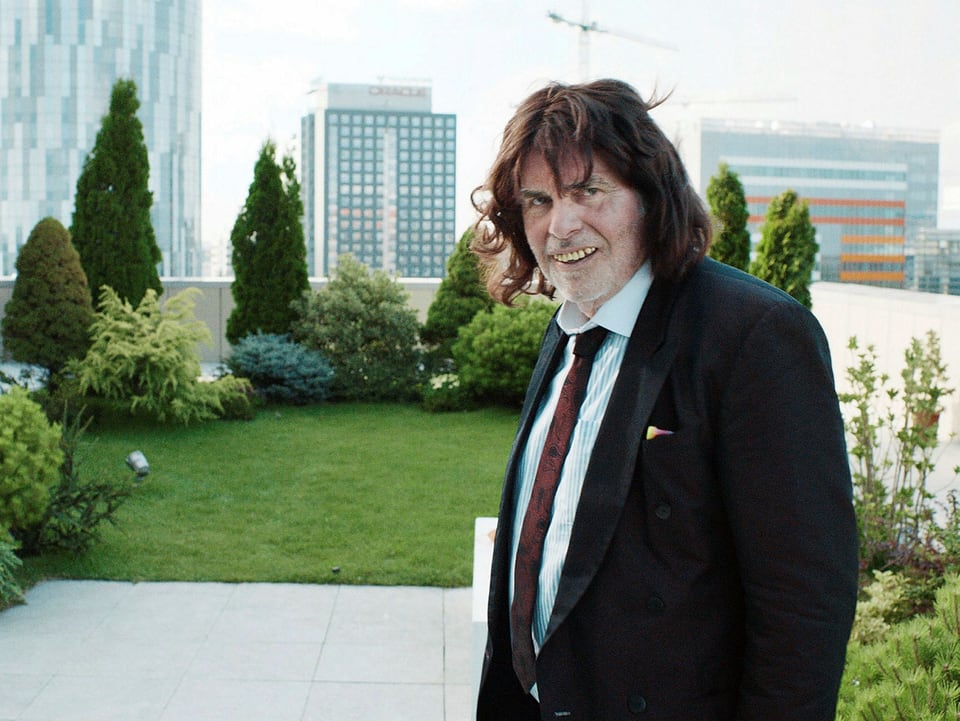 Toni Erdmann lächelt. Im Hintergrund sind Hochhäuser. 