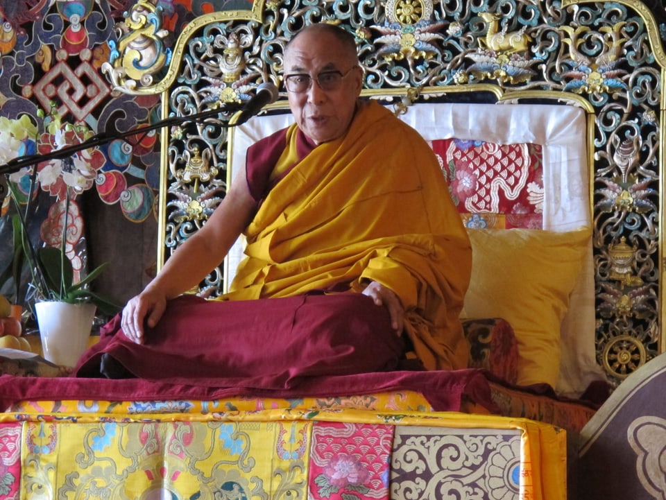 Der Dalai Lama sitzt im Schneidersitz auf einem verzierten Thron