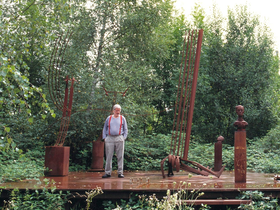 Der Künstler Schang Hutter gestreiftem Pulli und Hosenträgern steht neben seinen rostigen Skulpturen.