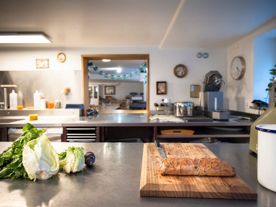 Eine Küche, auf der Ablage liegt ein Brett, ein Messer und Gemüse. Im Hintergrund: Geräte und weitere Räume.