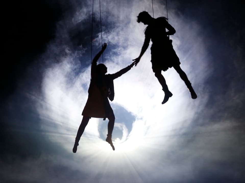 Im Gegenlicht: Ein Mann und eine Frau hängen an Seilen vom Himmel.