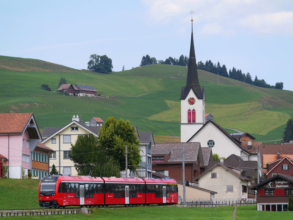 Drei Rote Zugwaggons der Appenzellerbahnen fahren gerade aus einem kleinen Dorf heraus.Im Hintergrund steht ein Kirchturm und ein Hügel.