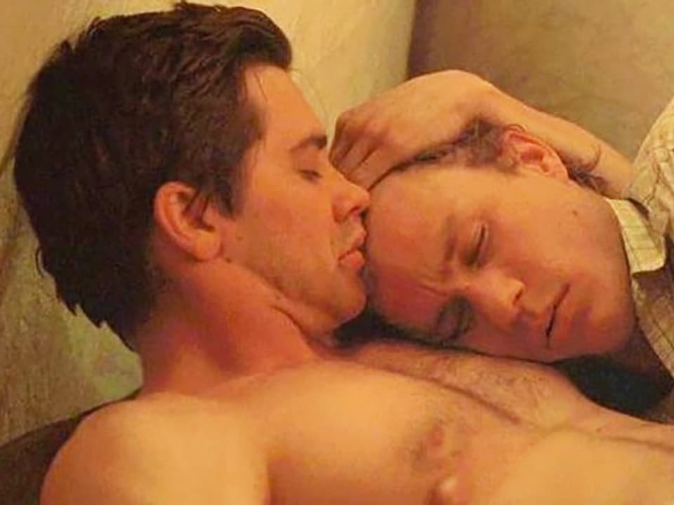 Zwei Männer sitzen eng beieinander, einer küsst den anderen auf die Stirn.