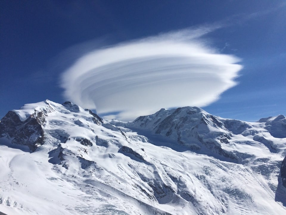 Im Vordergrund steht eine tiefverschneite Schneelandschaft. Über den Berggipfeln türmt sich eine Wolke auf, die einem Ufo gleicht. Innerhalb der Wolke kann man verschiedene Schichten erkennen, ähnlich einer Cremeschnitte. Die Schichten werden durch den Wind erzeugt.