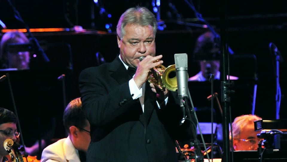 Ein Mann mit grauen Haaren spielt Trompete auf einer Bühne, im Hintergrund ein Orchester.