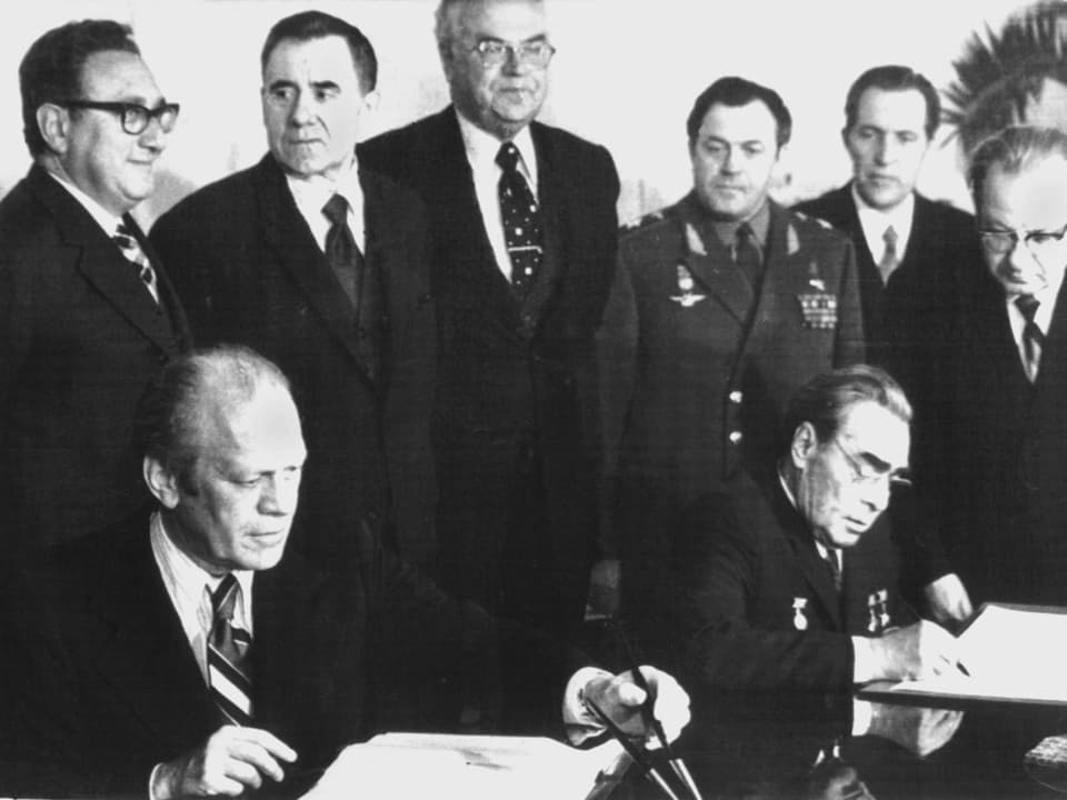 Zwei Männer sitzen und unterschreiben ein Abkommen, während sechs weitere Männer hinter ihnen stehen.