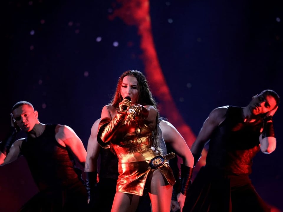Sängerin in goldenem Outfit performt auf der Bühne mit Tänzern und rotem Hintergrund.