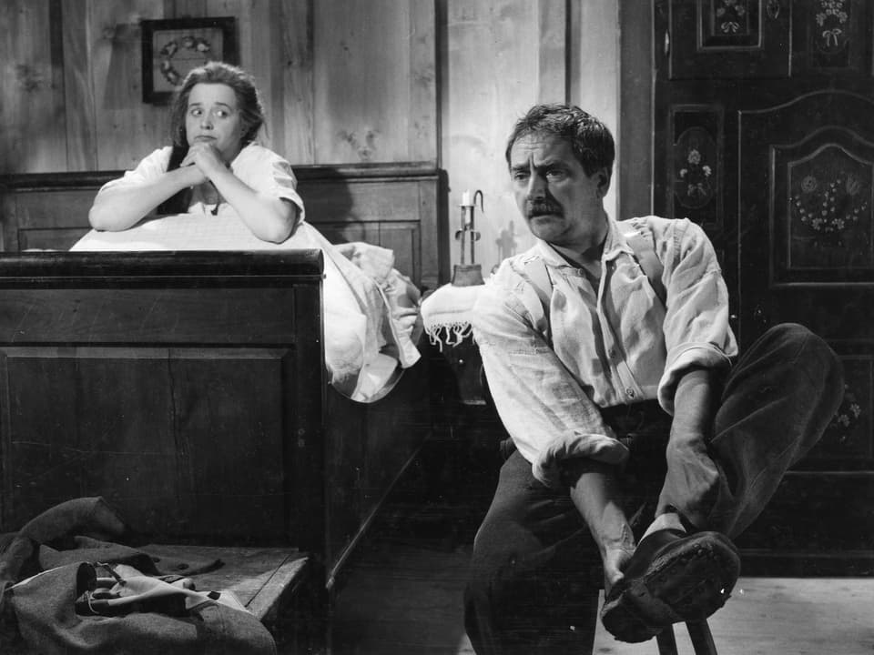 Ein Mann sitzt im Schlafzimmer auf einem Holzschemel und zieht sich die Schuhe aus. Hinter ihm im Bett sitzt eine Frau, die nachdenklich zu ihm schaut.