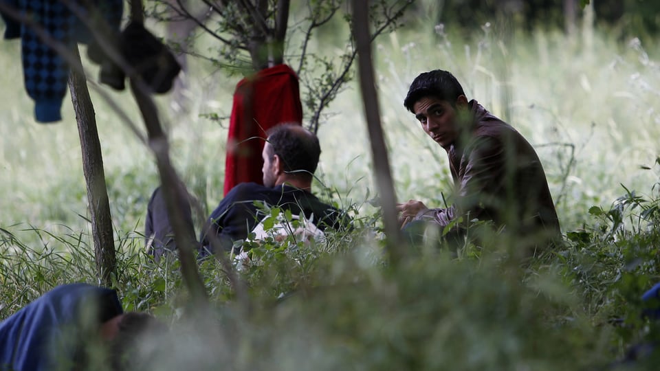 Zwei Flüchtlinge sitzen in einem Waldstück in Mazedonien, ihre nassen Kleider hängen an Ästen. 