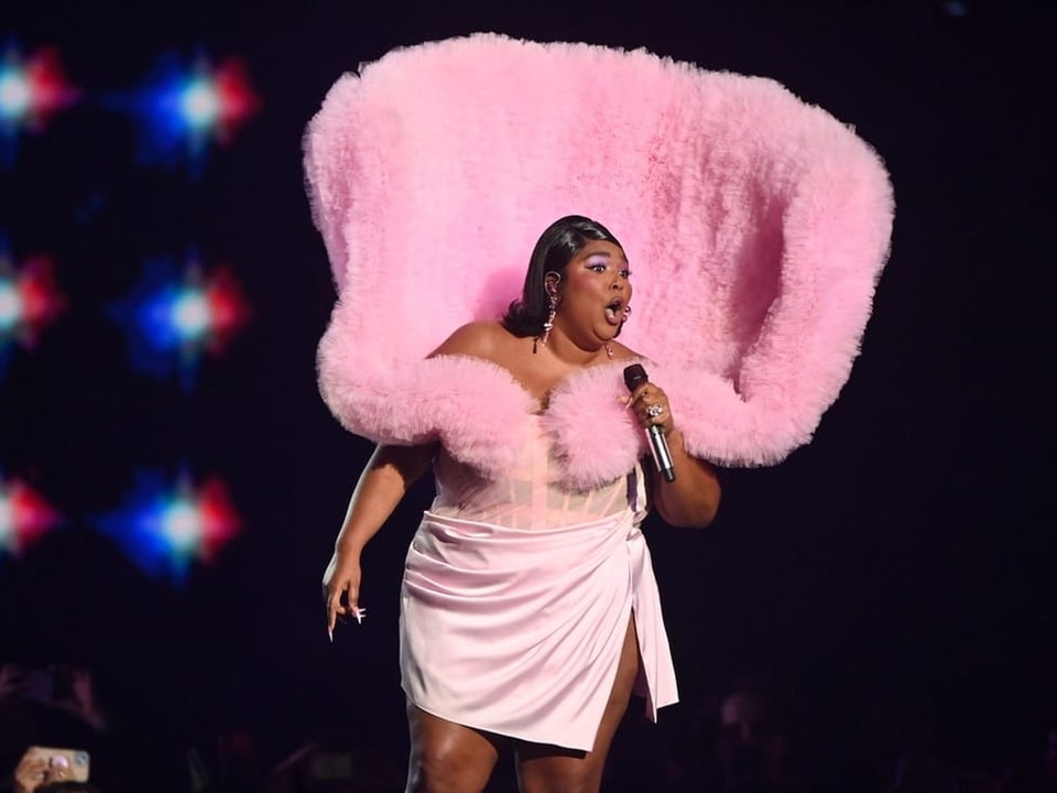 Eine Sängerin auf einer Bühne mit Mikrofon. Sie ist stark geschminkt und trägt ein Dress mit extrem hohem rosa Kragen.