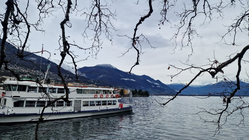 Blick auf den grauen Lago Maggiore mit einem Ausflugsschiff am Steg.