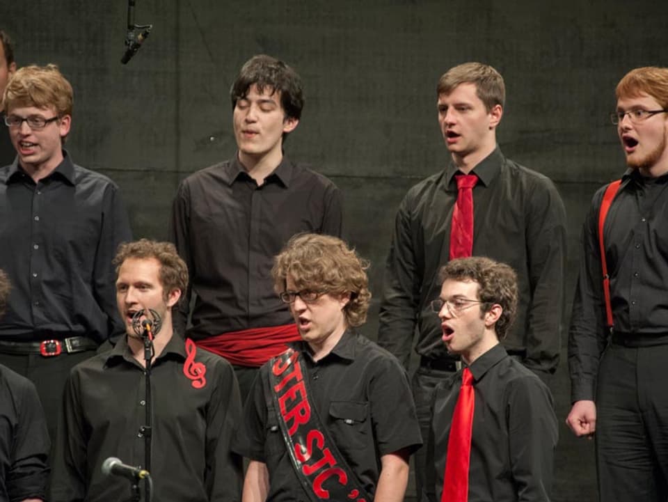 Die Sänger tragen schwarze Hemden und Hosen sowie rote Krawatten oder Schärpen.