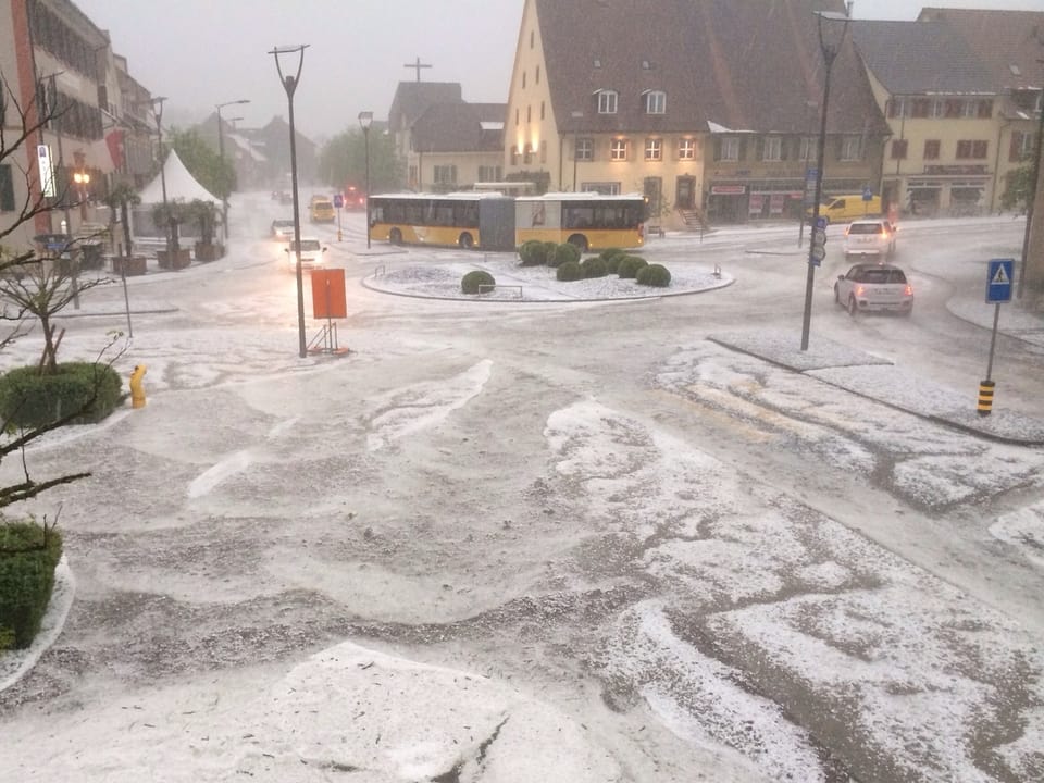 Ein Dorfplatz mit Bus und Autos, die Strassen sind weiss, voller kleiner Hagelkörner. 