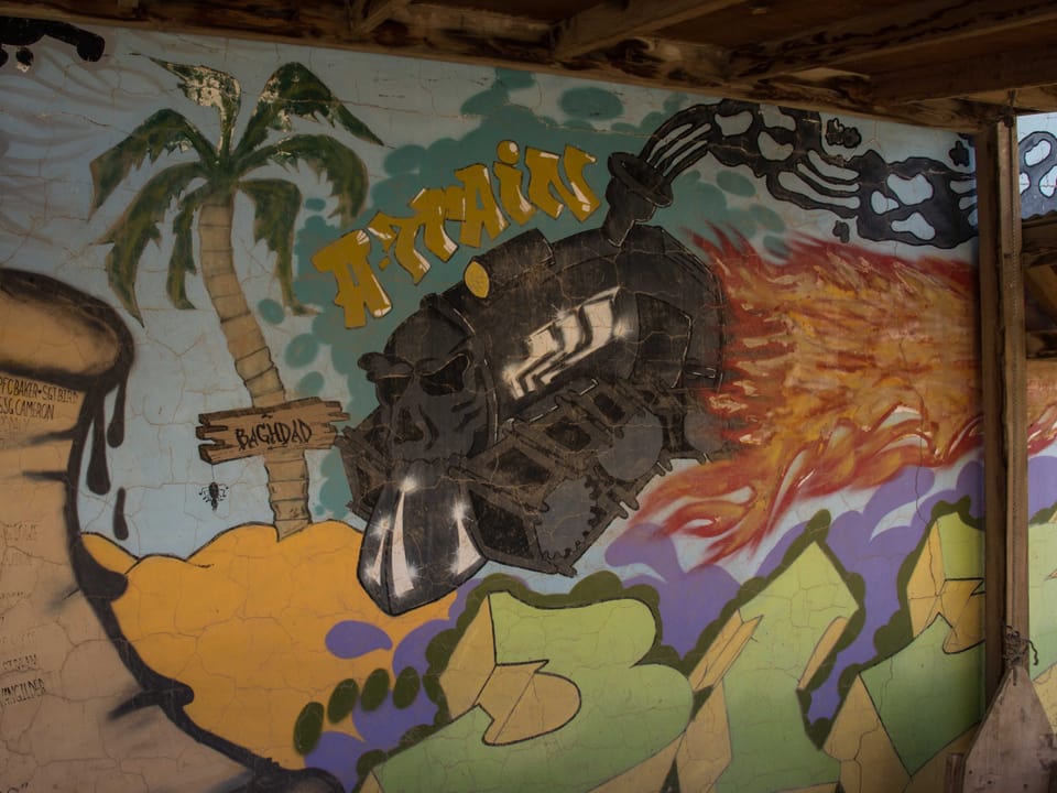 Ein Graffiti an einer Mauer, darauf zu sehen ist eine Palme mit dem Schild Bagdad und ein Zug der darauf zufährt.