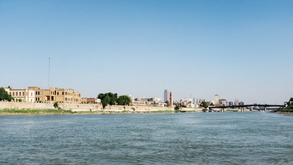 Wir sehen den Fluss Tigris. Der Himmel ist wolkenlos und blau, an der linken Uferseite sind Gebäude erkennbar.