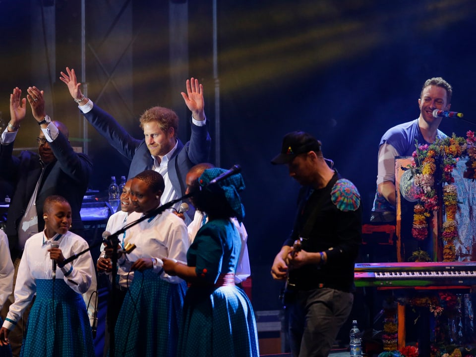 Prinz Harry steht auf der Bühne und hält die Hände hoch. Im Hintergrund sitzt Chris Martin von Coldplay am Piano.