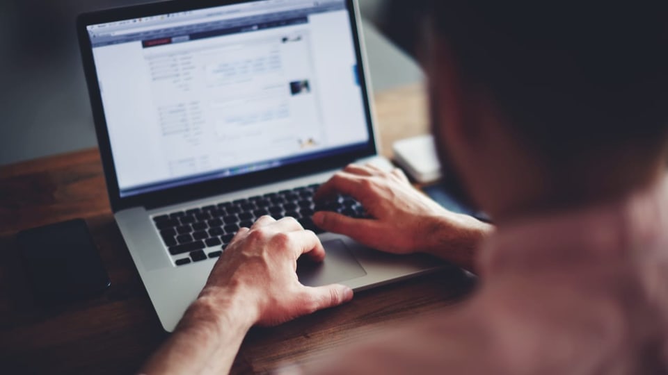 Junger Mann sitzt an einem Holztisch und tippt in seinen Laptop.