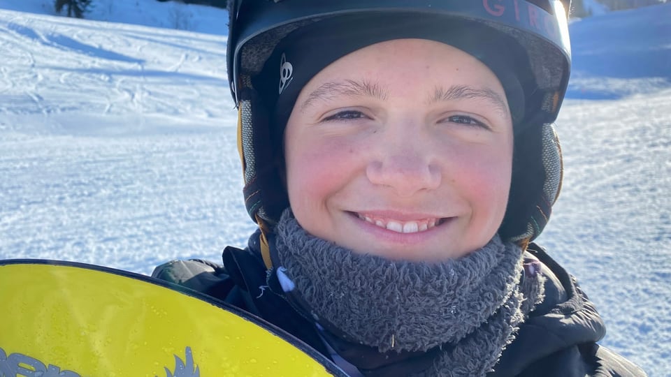Gaël mit Snowboard auf der Piste und lächelt in die Kamera.