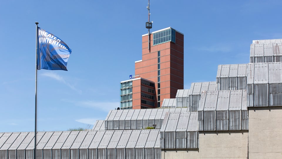 Das graue Dach des Theater Winterthur, davor eine blaue Fahne, dahinter ein Turm.
