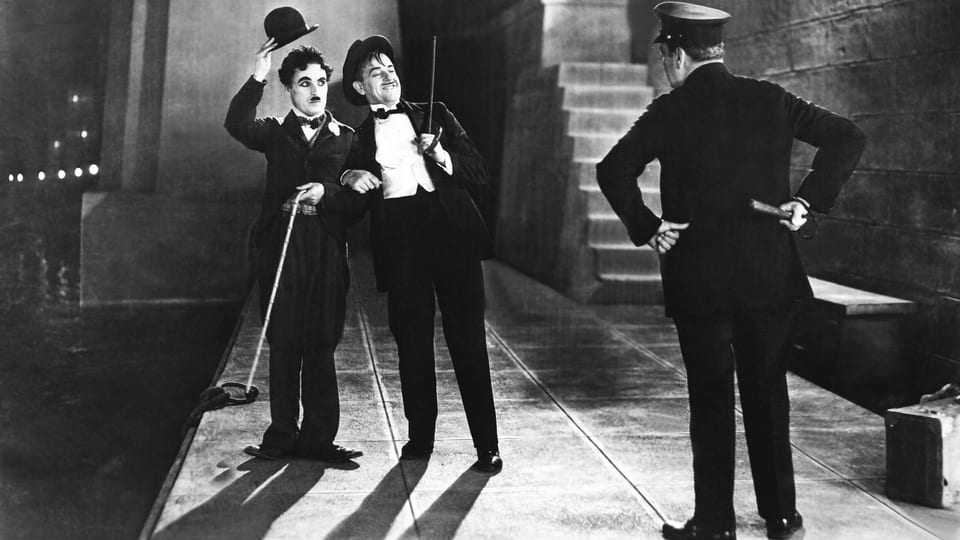 Schwarzweiss-Filmszene: Chaplin mit Melone uns STock grüsst zusammen mit einem betrunken aussehneden Mann einen Polizisten.