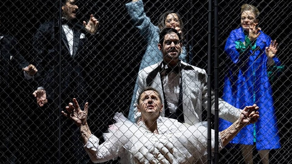 blonder Mann an Gitterwand, verzweifelt die Hände am Gitter, dahinter vier fröhliche Menschen.