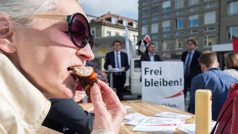 Eine Frau beisst in eine Bratwurst, im Hintergrund stehen SVP-Wahlkampfleiter.