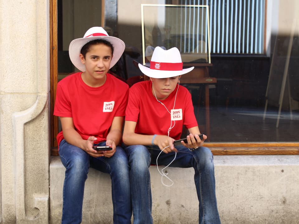 Zwei Knaben roten T-Shirts und rot-weissen Hüten mit Smartphone in der Hand, sitzen auf Schaufensterbank.